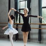 Full body of Asian teacher in black outfit and pointe shoes holding hand of female student while practicing dance movement